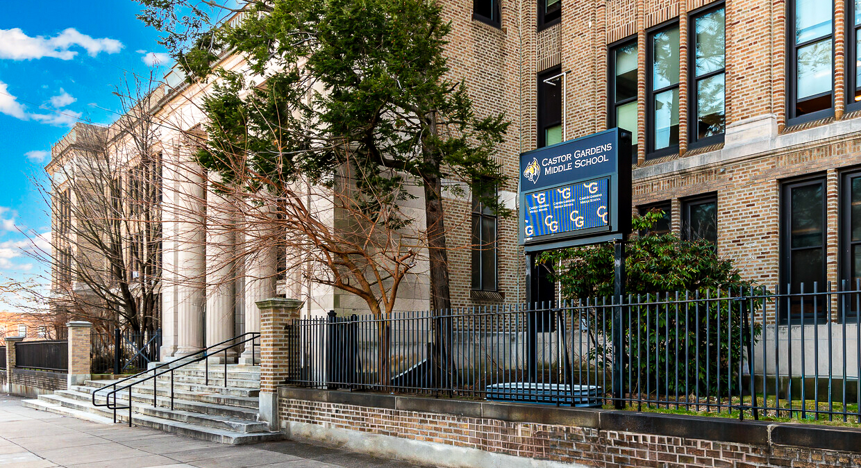 Exterior view of Castor Gardens Middle School building with brick facade, front steps, black iron fence, and school sign displaying the school name.